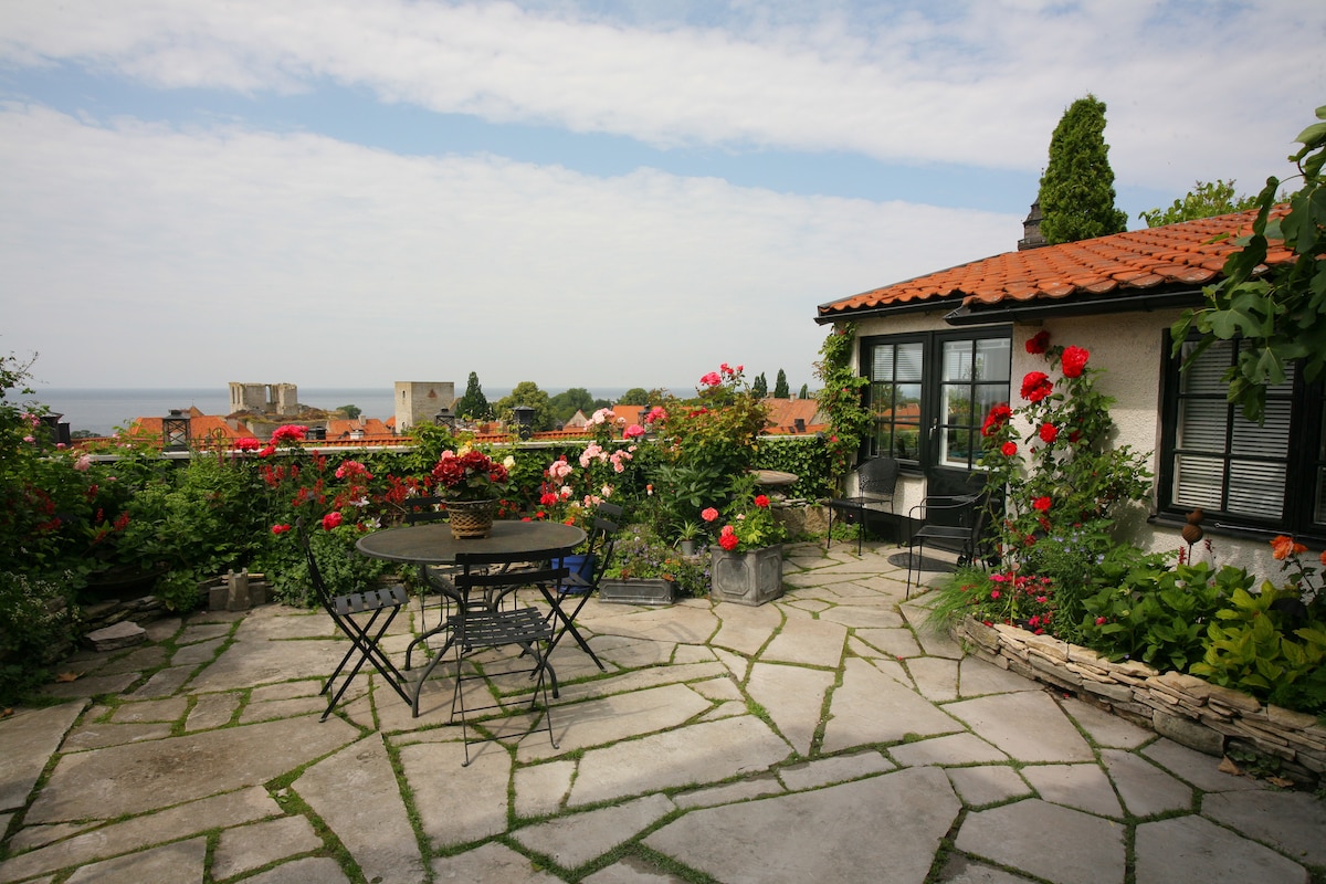 A terrace is surrounded by a colorful array of blooming roses, with stone pathways leading to a cozy seating area. A round table and chairs are positioned for relaxation, offering a view of the distant sea and nearby rooftops under a partly cloudy sky.