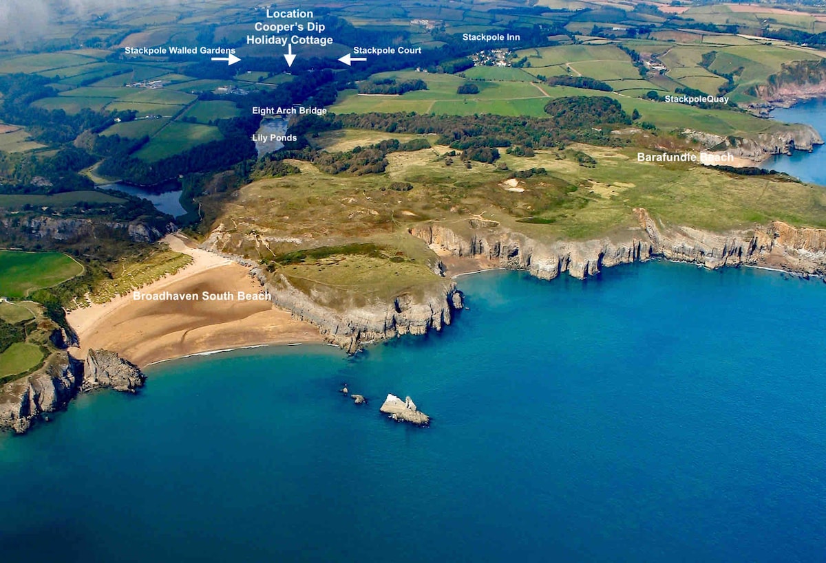 An aerial view captures the stunning coastline of Pembrokeshire, featuring Broadhaven South Beach and the surrounding cliffs. The tranquil blue waters are contrasted by lush green fields and wooded areas, with several notable landmarks including the Stackpole Walled Gardens and the picturesque Stackpole Inn.
