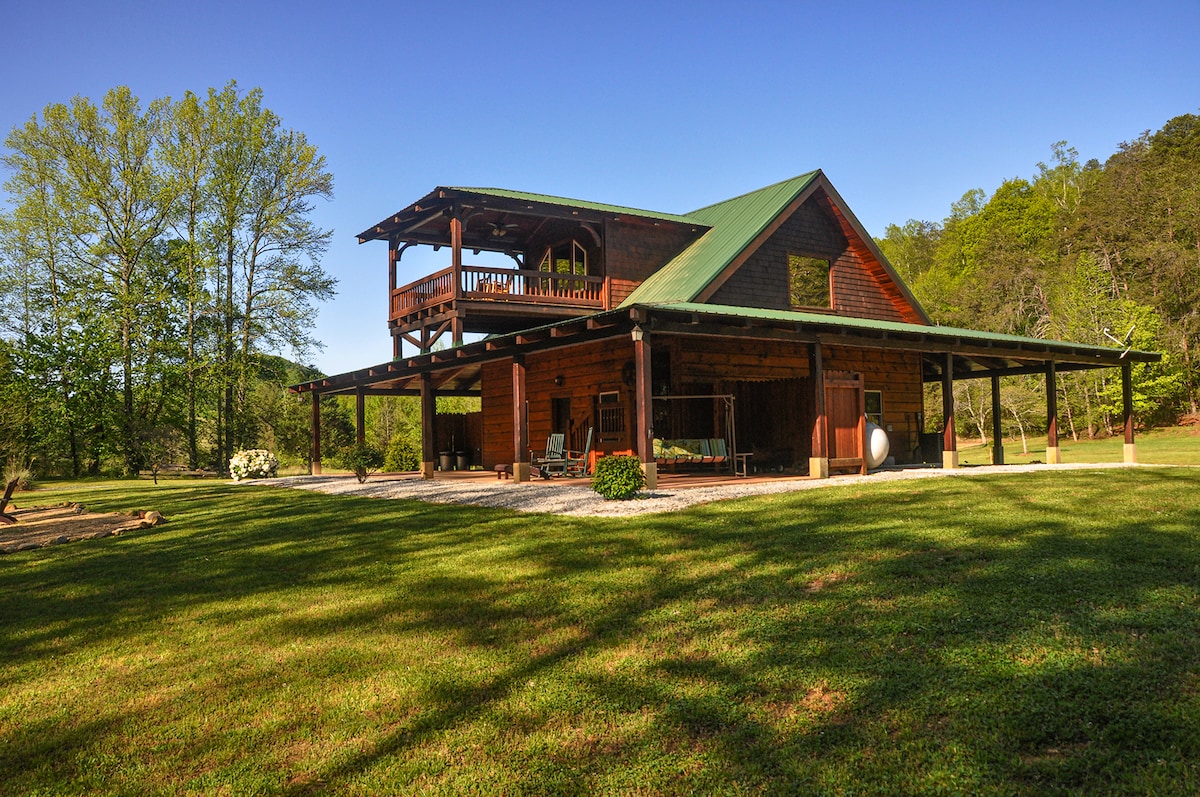 The exterior showcases a two-story cabin with a rustic wooden facade and green metal roof. A spacious covered porch extends from the lower level, while a large upper deck offers views of the surrounding greenery. Lush grass surrounds the cabin, enhancing its secluded feel.