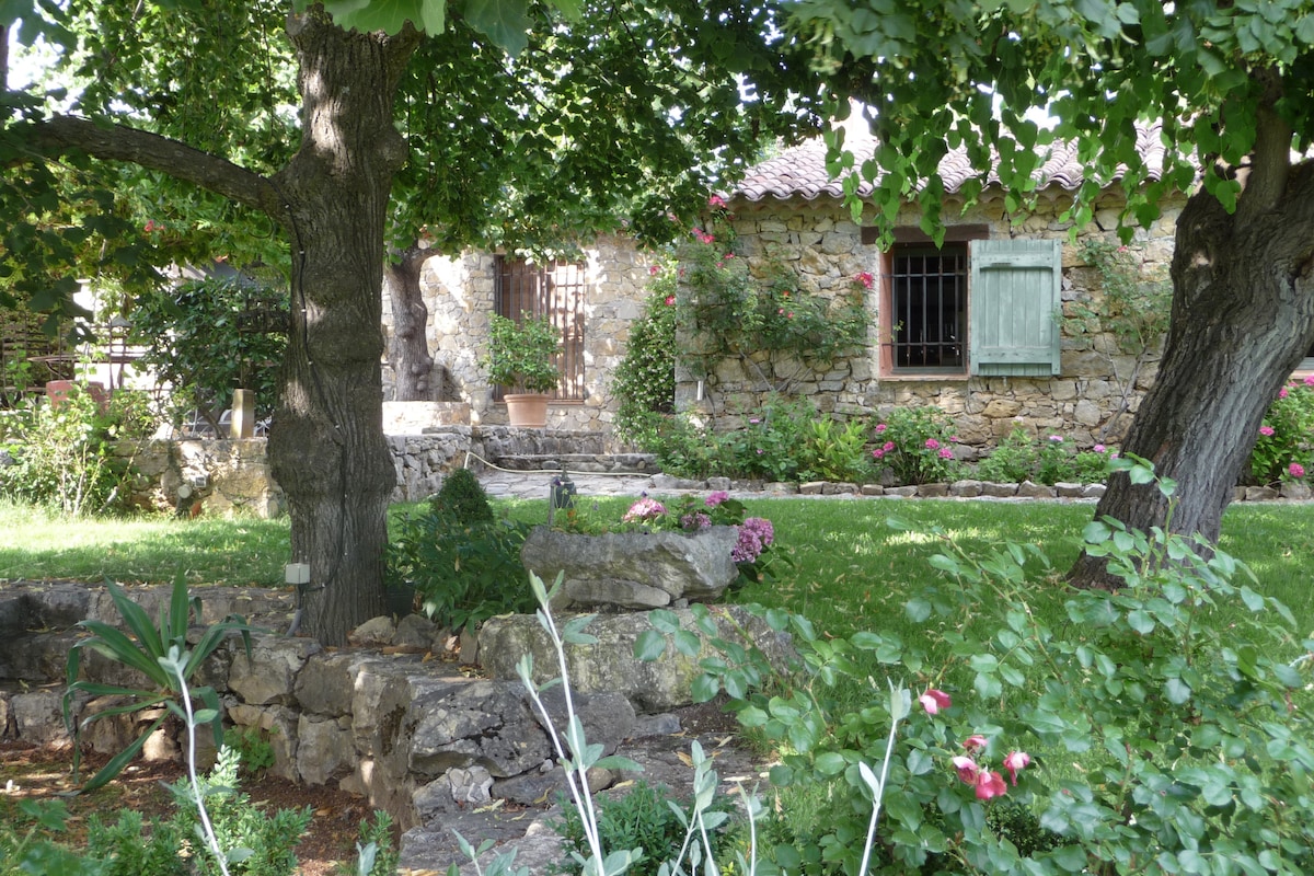 A stone cottage stands in a lush garden surrounded by flowering plants and trees. The rustic architecture features wooden shutters and a pathway leading to the entrance. A variety of greenery is visible, offering a natural, tranquil setting.