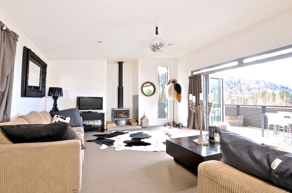 A modern living area features a light-colored sofa and a dark coffee table, accented by a decorative cowhide rug. A fireplace is positioned against a wall, and large glass doors open to a sunlit deck, depicting an indoor outdoor flow.