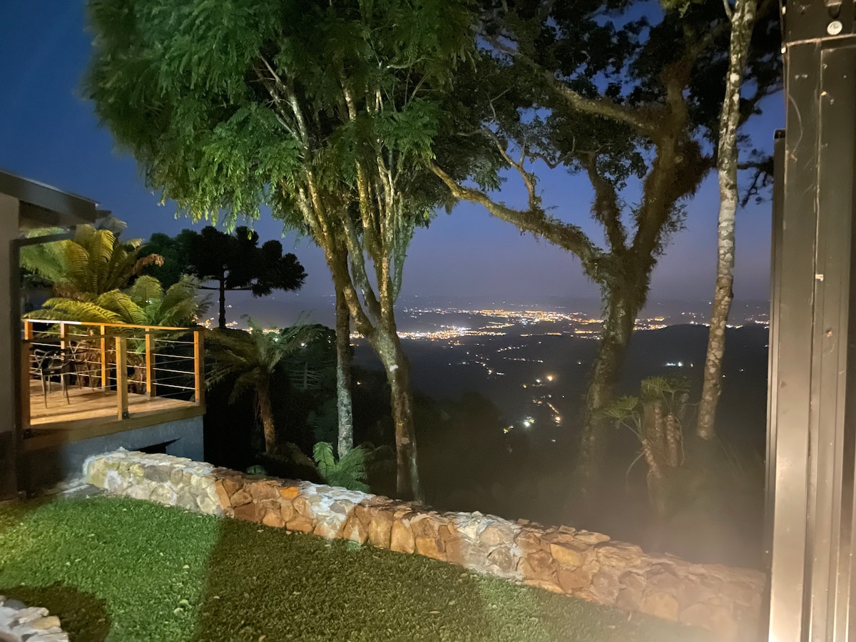 A tranquil nighttime scene reveals a panoramic view of illuminated valleys below. The landscape is framed by lush trees, creating a serene atmosphere. In the foreground, a stone wall complements the green grass, while the distant city lights twinkle in the background.