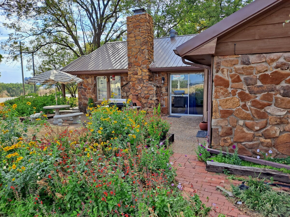 The exterior showcases the historic stone façade of the Old Station Main House, surrounded by vibrant flowerbeds. A shaded picnic table sits on a gravel area, with a patio area visible through large glass doors, offering a glimpse of the interior and scenic landscape beyond.