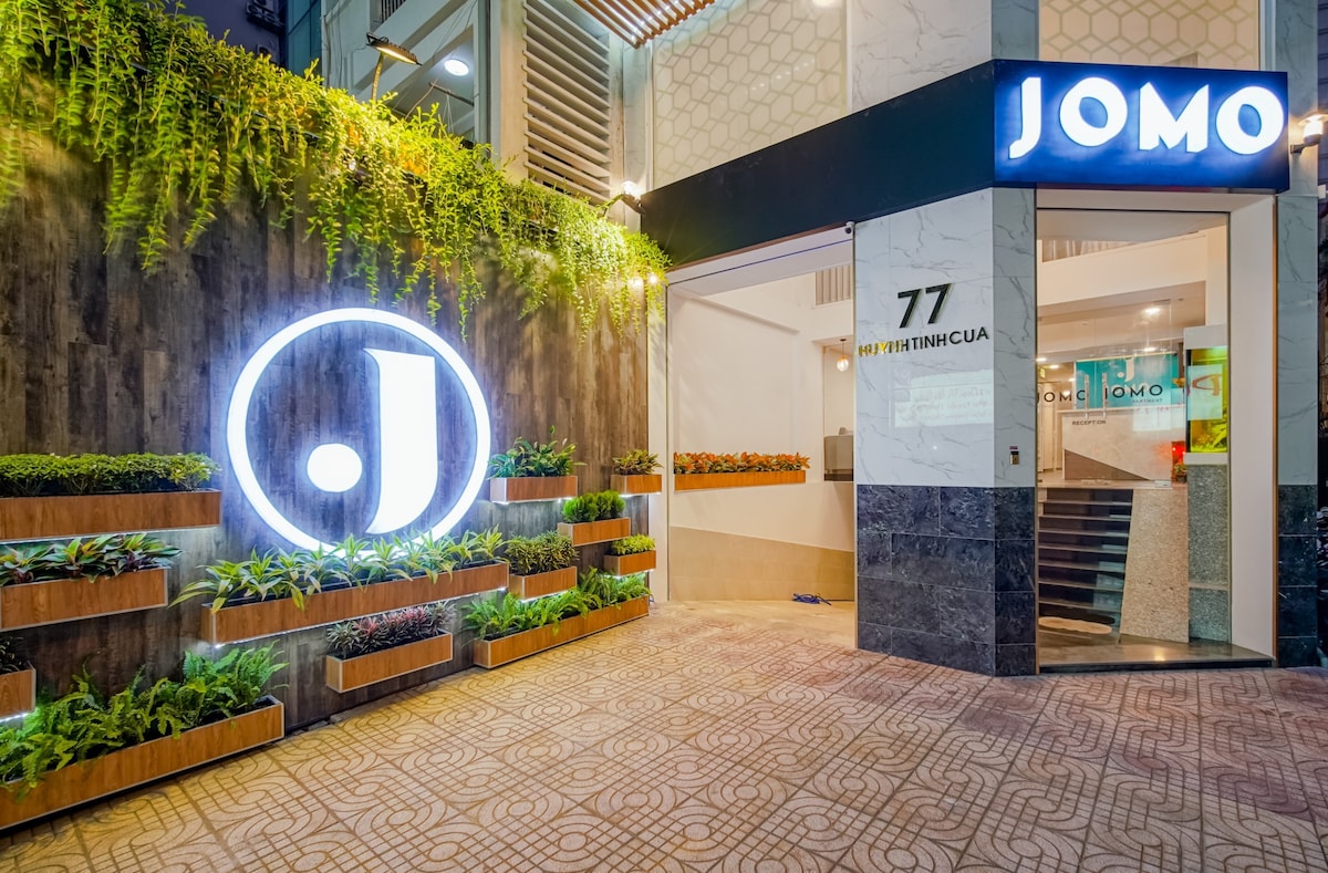 The entrance to JOMO features a modern facade with a prominent illuminated logo. Lush greenery is displayed in wooden planters along the wall, creating a welcoming atmosphere. The tiled pathway leads to a stylish reception area, marked by the building number prominently displayed.