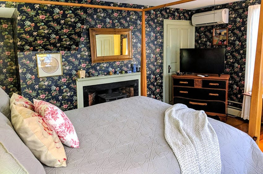 The guest room features bold floral wallpaper and a spacious queen-size four-poster bed with decorative pillows. A large flat-screen TV is positioned on a wooden dresser, and a mirror above the fireplace reflects warm tones. Natural light enters through a nearby window.