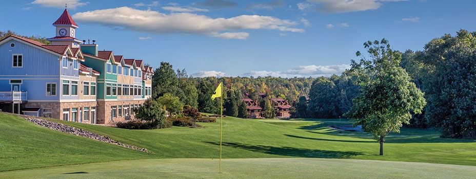 A well-maintained golf course is depicted, featuring a vibrant green expanse and a yellow flag marking the hole. The resort building stands in the background, showcasing multiple stories and a peaked roof, surrounded by trees and colorful foliage.