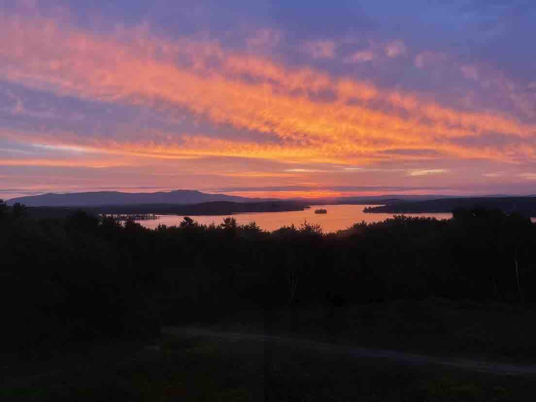 A vibrant sunset casts shades of pink and orange across the sky, reflecting on the water below. Silhouettes of distant mountains and trees frame the serene lake, creating a peaceful natural scene.