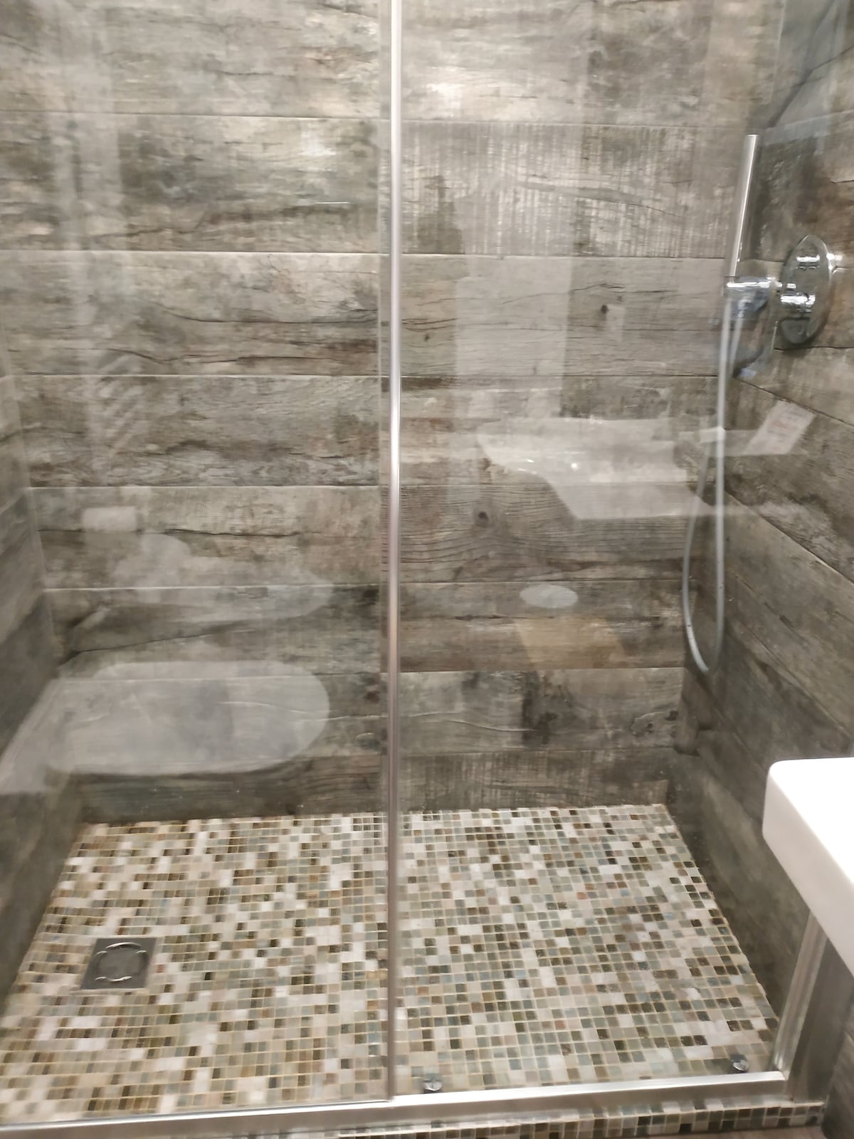 A modern shower area features transparent glass walls, revealing a spacious shower with a rainfall showerhead. The floor is adorned with mosaic tiles in neutral tones, while natural wood-like wall panels create a contemporary aesthetic.