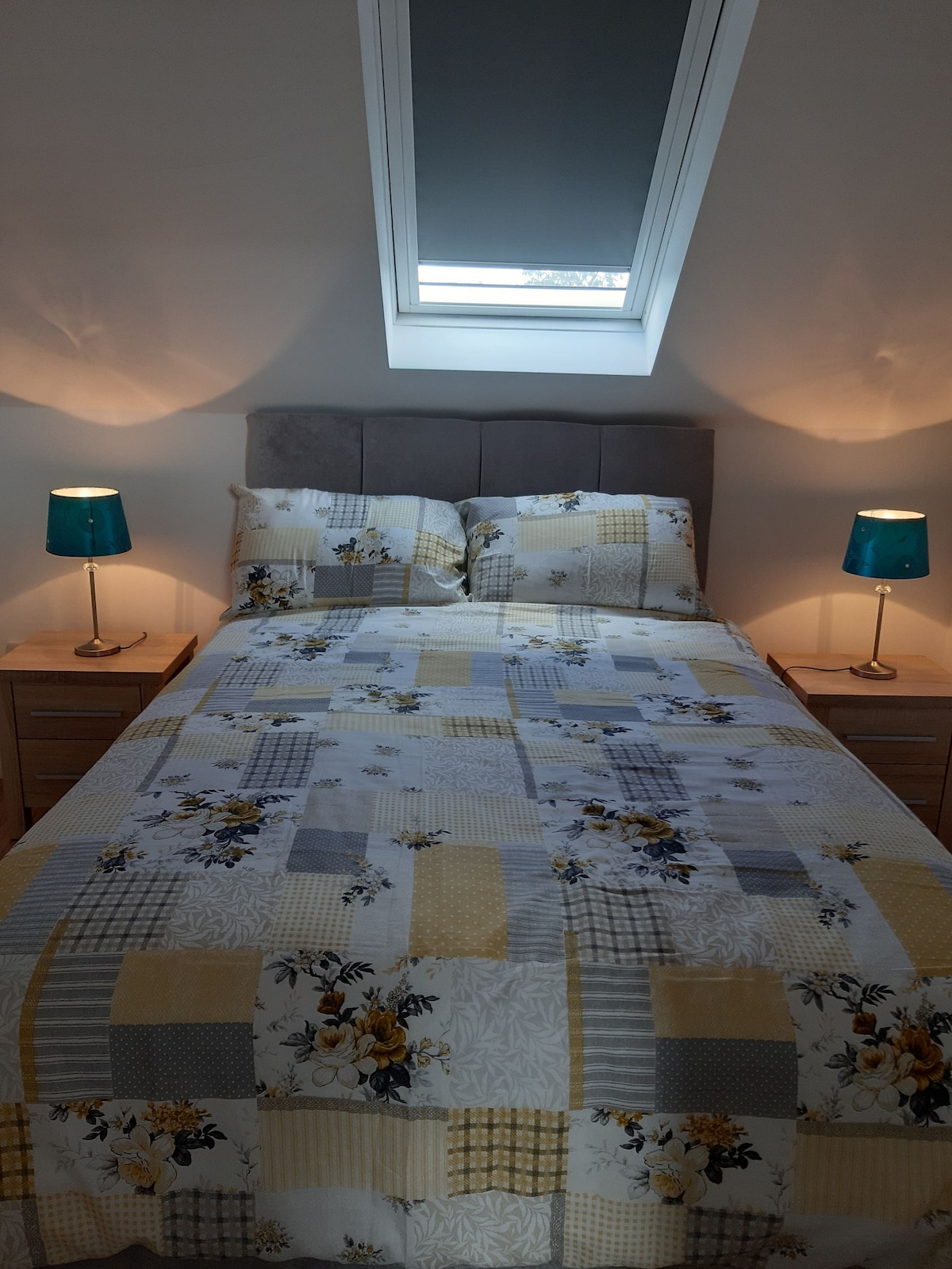A double bed is dressed in a patchwork quilt featuring floral and geometric patterns in yellow, gray, and white tones. Two bedside tables are positioned on either side of the bed, each topped with teal lamps. Natural light enters through a skylight above.