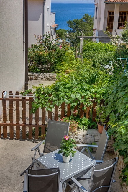 An outdoor area features a circular table surrounded by four chairs. A small potted plant sits at the center of the table. Lush greenery lines the space, leading towards a scenic view of the sea in the background.