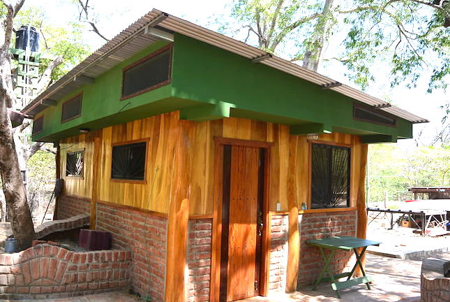 The exterior of a charming, rustic house features natural wood siding with green accents, blending into the surrounding trees. A sturdy wooden door and multiple windows provide light and ventilation. A small table is positioned outside, adding to the welcoming feel of the property.