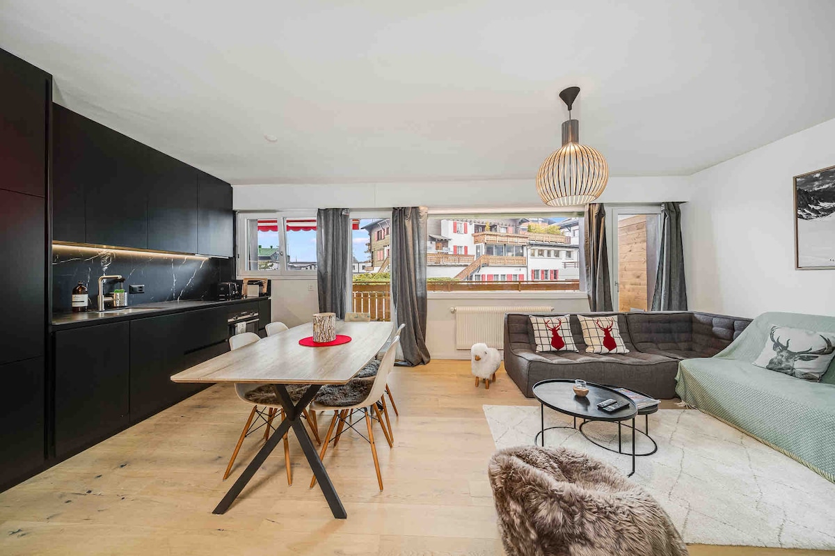 An open living area features a modern kitchen with dark cabinetry and a dining table for six. Large windows allow natural light, offering views of the surroundings. A comfortable gray sofa and coffee table are centered on a light area rug, providing a cozy space.
