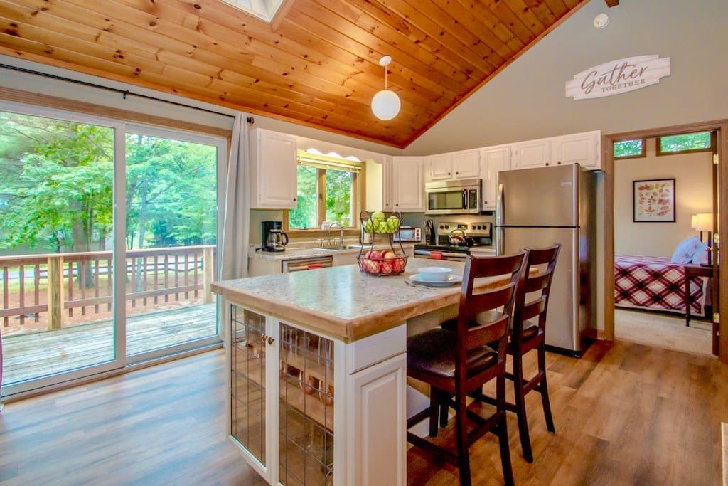 A well-lit kitchen features white cabinetry and a center island with seating for two. Large glass doors open to a deck, allowing natural light to fill the space. Pine wood ceilings add warmth, and modern appliances are visible, complementing the inviting atmosphere.