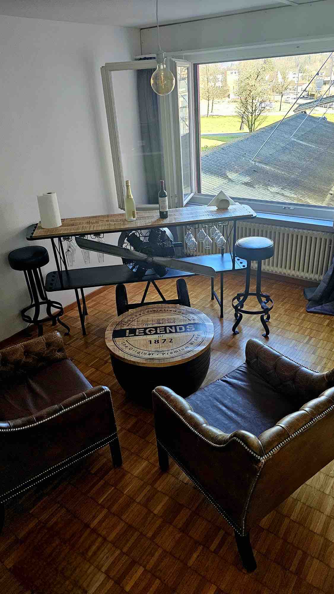 A cozy seating area features two brown leather chairs and a circular coffee table. A tall, sleek bar table is accompanied by two high stools. Natural light floods the space through a large window, creating a bright and airy atmosphere.