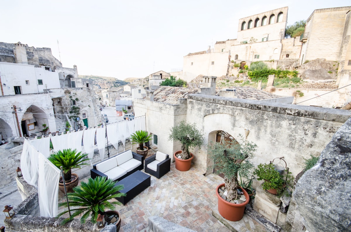 A private terrace is presented with outdoor seating arrangements, featuring a sectional sofa and potted greenery. The space offers views of the surrounding ancient structures and landscape, highlighting the unique architecture of Matera's Sassi.