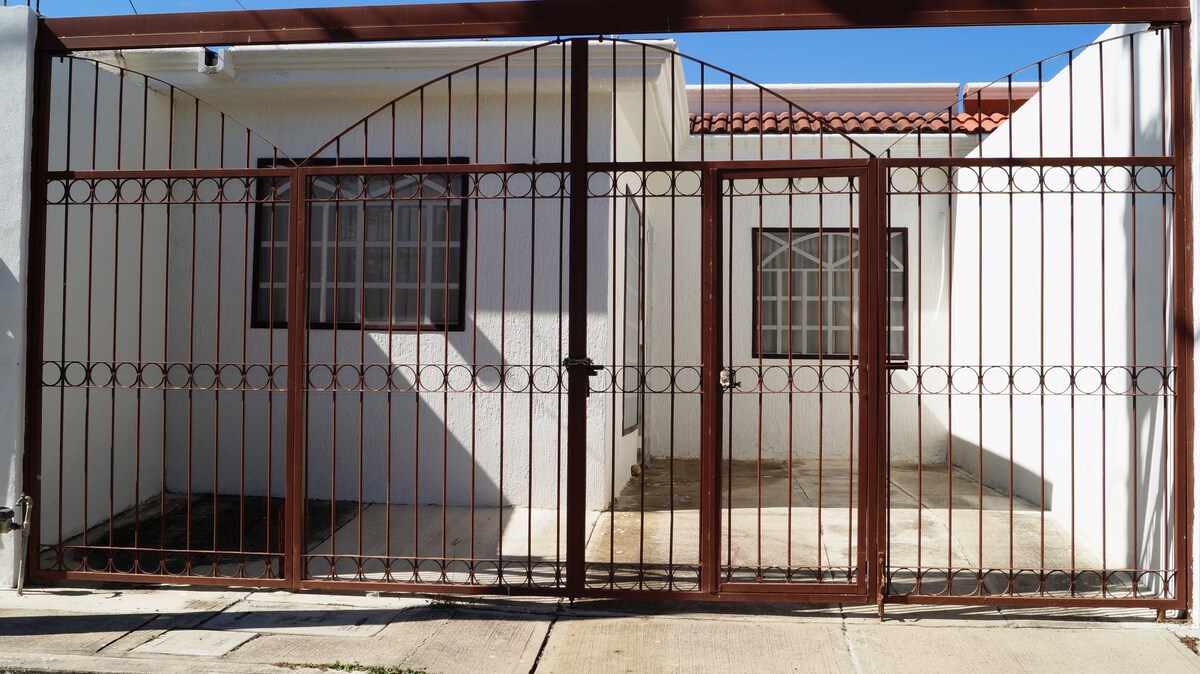 A gated entrance presents an iron fence with decorative detailing, leading to a simple, single-story home. Two windows are visible on the left side, allowing natural light into the living space. A tiled pathway extends towards the entrance, framed by a clear sky.