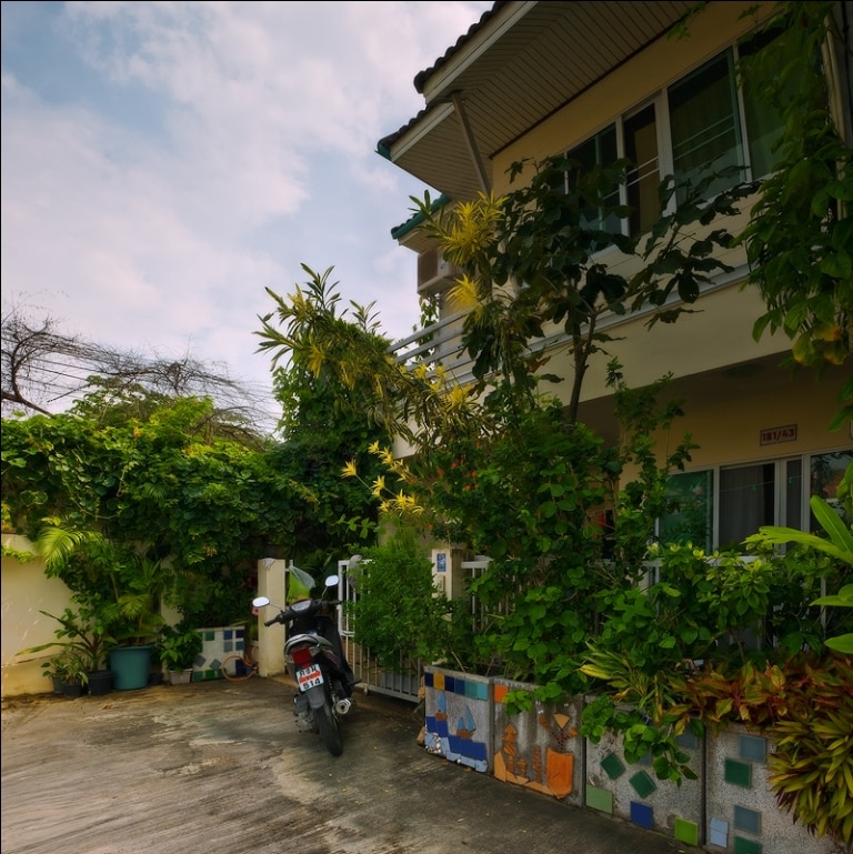 A two-story townhouse is surrounded by lush greenery, featuring vibrant plants and colorful tiles along the pathway. A motorcycle is parked at the entrance, with a small outdoor area that offers a welcoming and private ambiance.