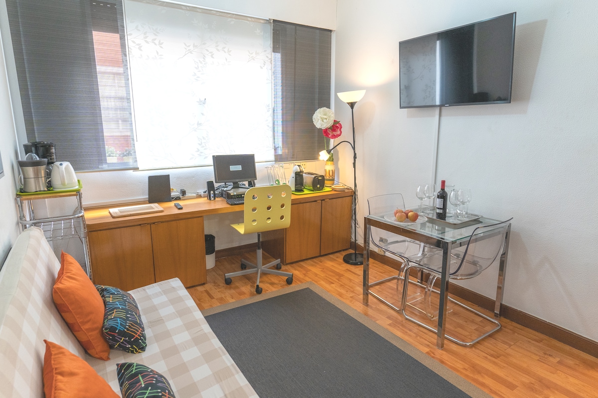 A well-organized living space features a comfortable sofa bed and a small dining area with a glass table and four chairs. A work desk with a computer is provided, alongside a wall-mounted television. Natural light enters through the window, enhancing the atmosphere.