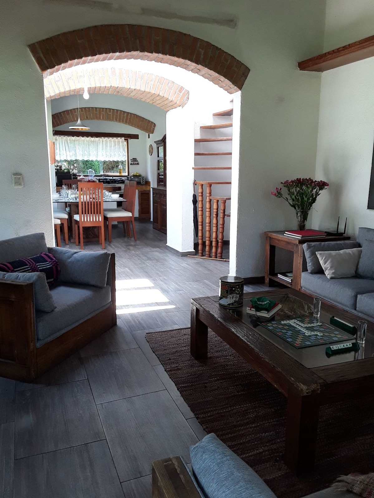 A welcoming living area is viewed from a relaxed seating arrangement. Natural light filters through a nearby window, highlighting the wooden beams and arches that frame the space. The adjacent dining area is visible, with a wooden table set for meals, and a staircase leading to an upper level.