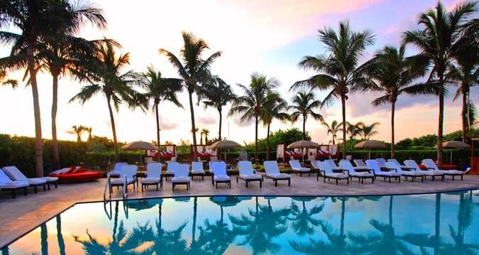 A serene pool area is surrounded by tall palm trees, with lounge chairs arranged neatly along the edge. The water reflects the vibrant colors of the sunset, creating a tranquil atmosphere. Umbrellas provide shade for relaxation.