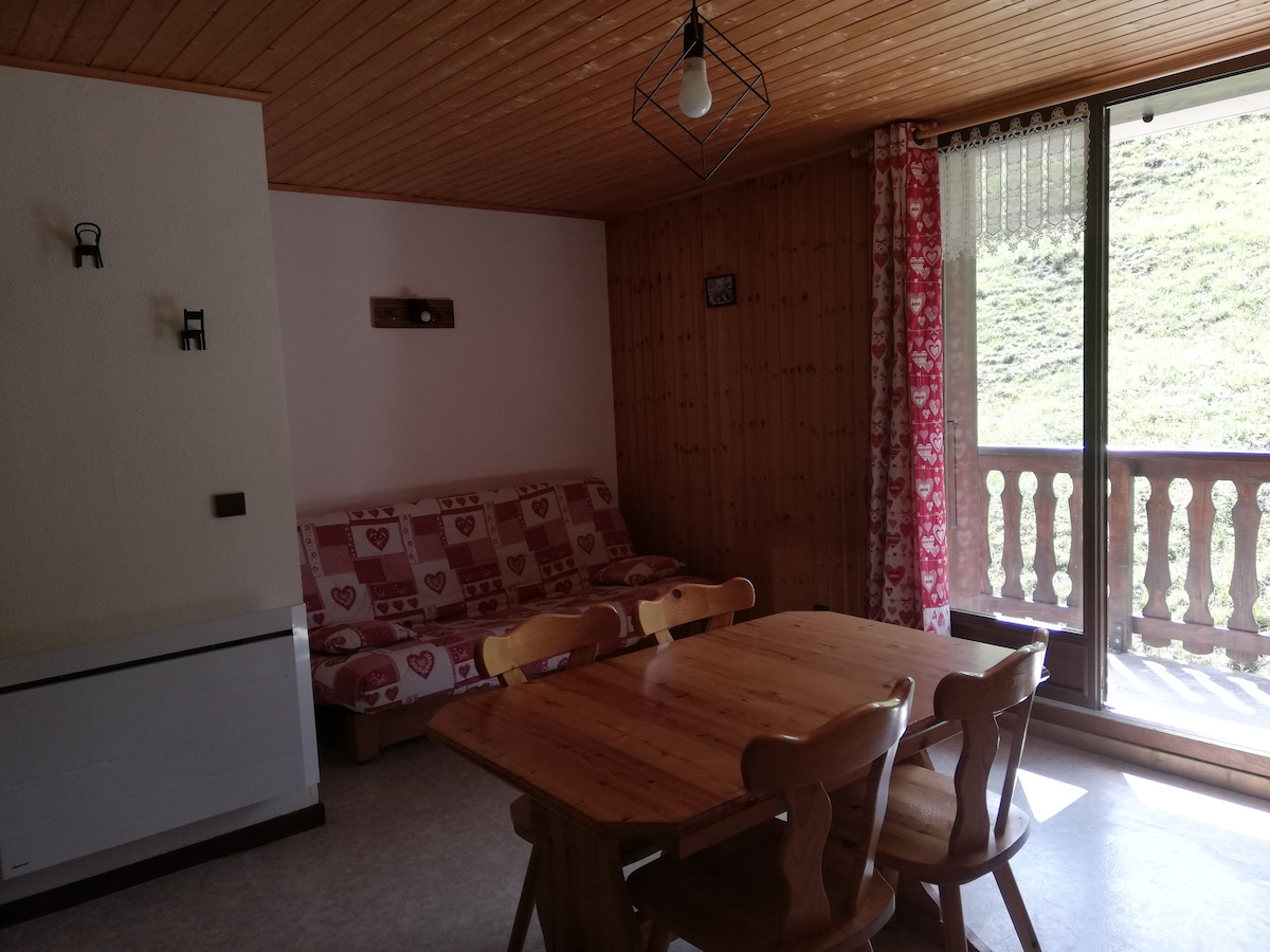 A living area features a red-patterned sofa that converts into a bed, positioned near a wooden dining table with four chairs. Large glass doors open to a balcony, allowing natural light to fill the space. The walls have a warm wooden finish.