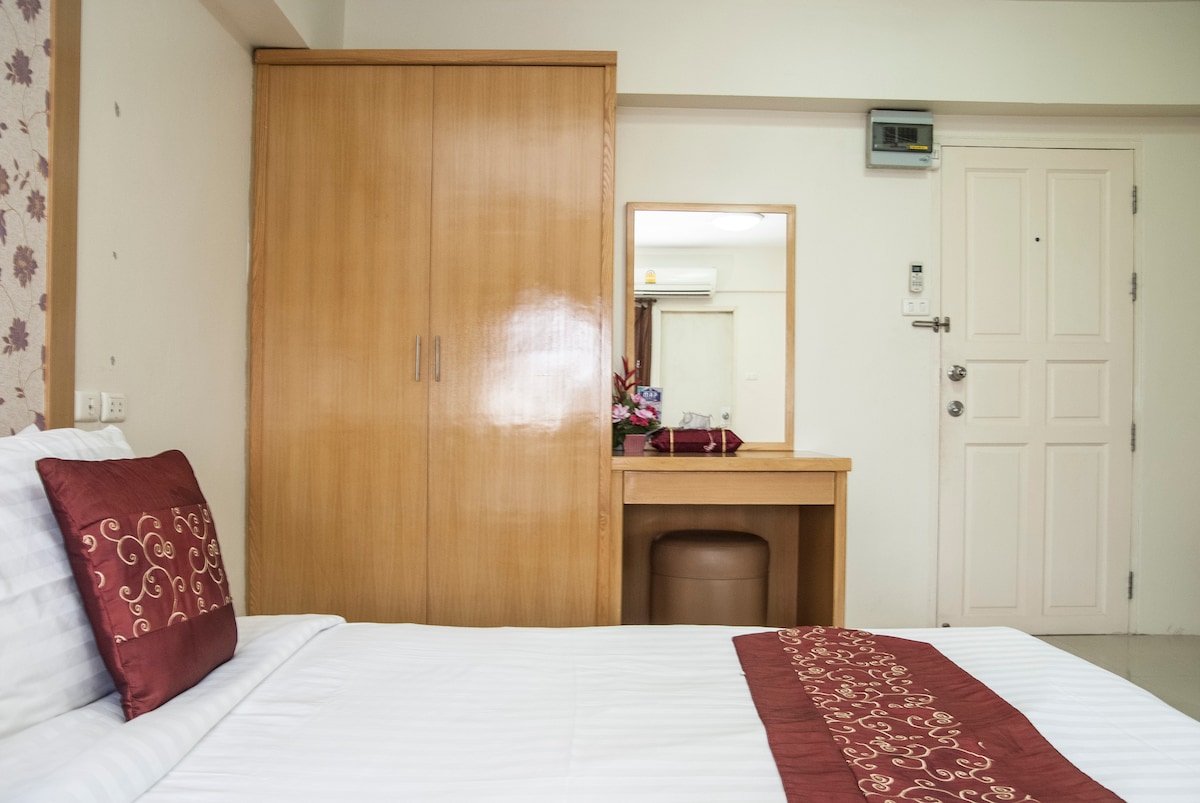 A well-organized Deluxe Studio room features a spacious bed adorned with decorative pillows and a coordinating bed runner. A wooden wardrobe and a mirrored vanity with a comfortable stool are visible, while soft natural light illuminates the serene space.