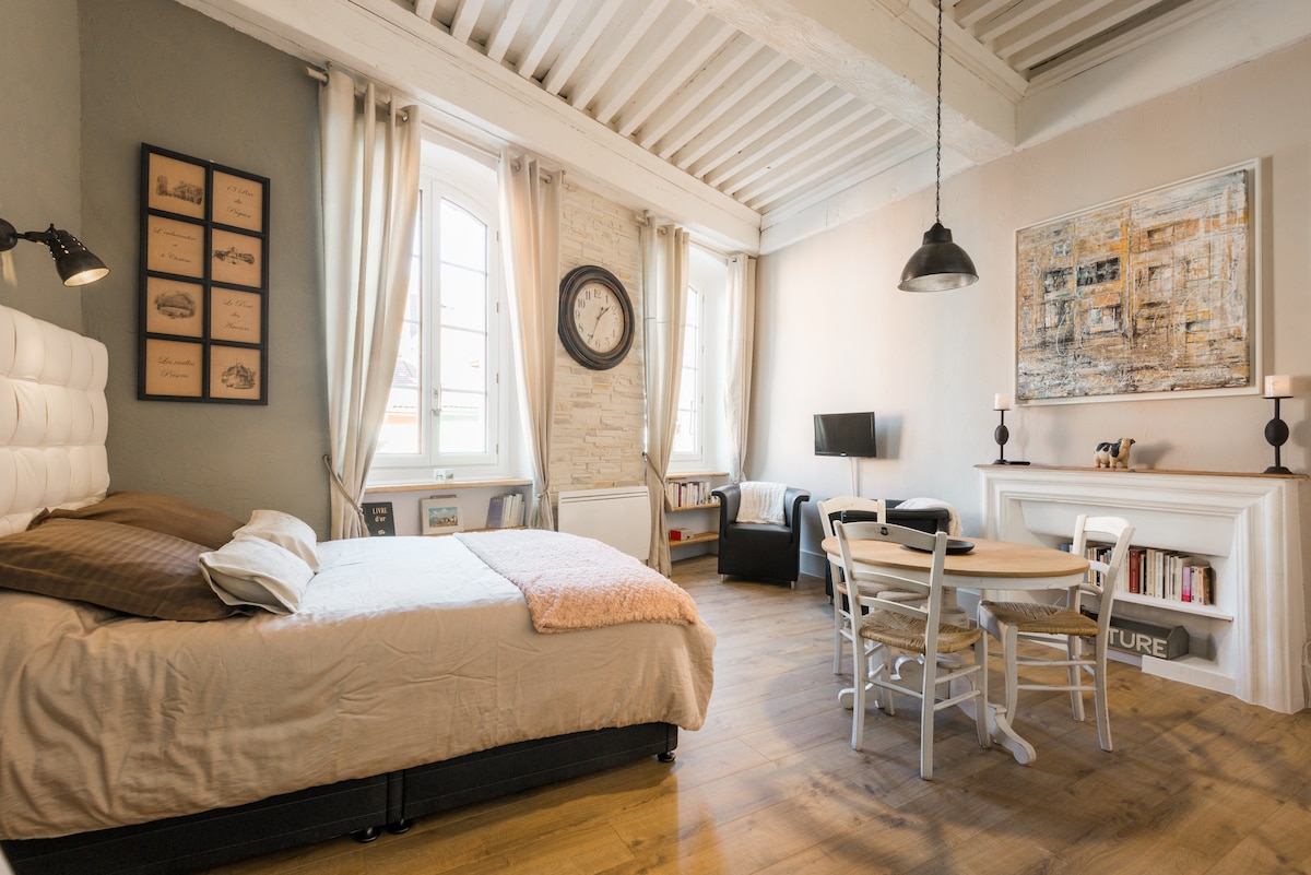 A cozy studio space features a spacious bed dressed in soft linens. Natural light filters through large windows, highlighting the warm wood flooring and inviting sitting area. A round dining table complements the decor, with framed artwork and decorative elements positioned throughout the room.