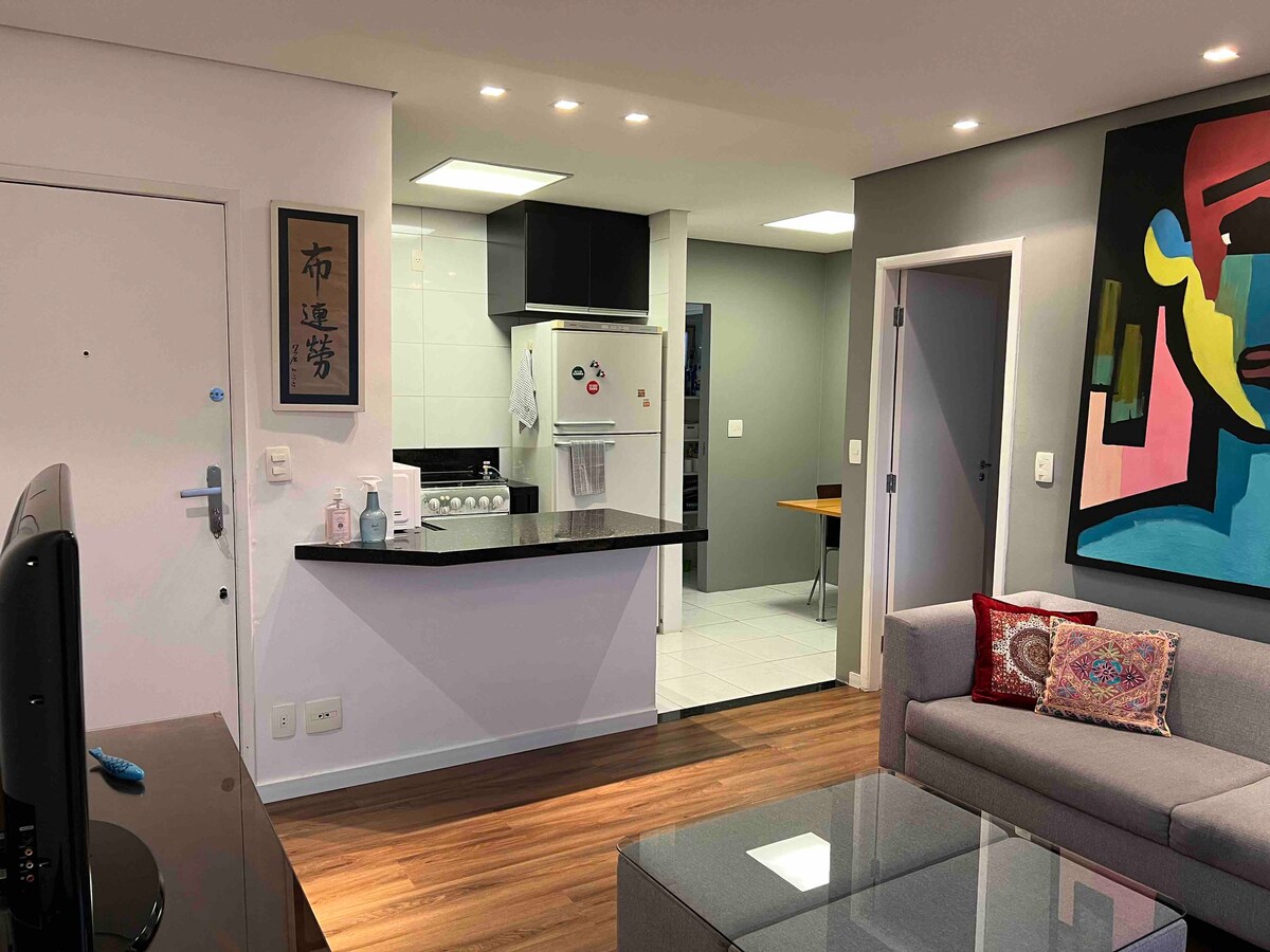 The living area features a modern sofa with decorative cushions, a glass coffee table, and a TV. An open kitchen is visible in the background, equipped with a stove and refrigerator. The walls display neutral tones with artwork enhancing the space.