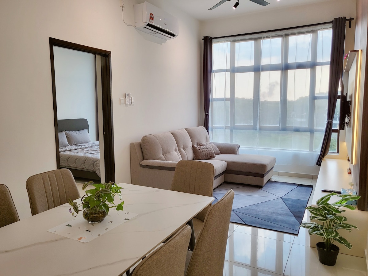 A light-filled living area is present, featuring a comfortable grey sectional sofa positioned next to a large window with sheer curtains. A neutral-toned dining table with matching chairs is seen in the foreground, complemented by a small potted plant. The room offers a clean, modern aesthetic.