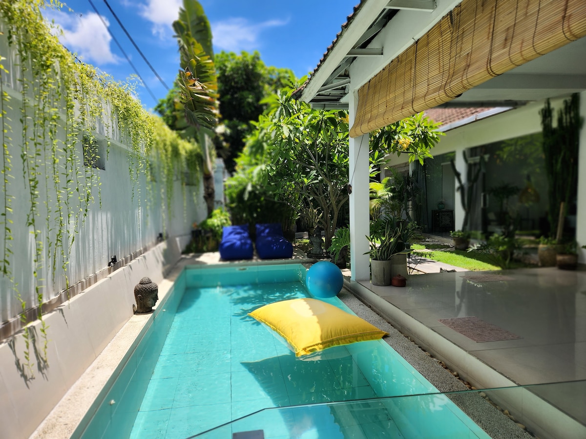 A serene pool area is bordered by lush greenery and bright blue bean bag chairs. A vibrant yellow cushion floats on the water, while sunlight filters through leafy trees, creating a peaceful environment for relaxation.