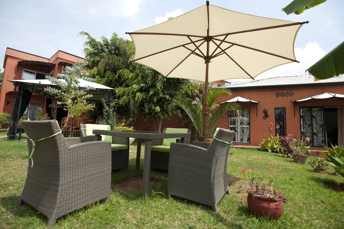 A well-kept garden area features a round table surrounded by comfortable chairs under a large umbrella. Lush greenery and various plants are visible in the vicinity, enhancing the outdoor relaxation space. The building in the background showcases large windows with a tranquil setting.
