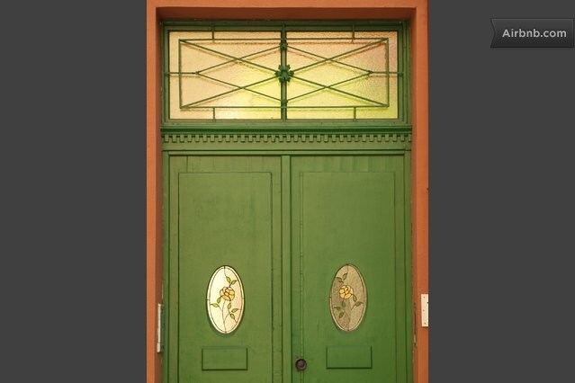 A green double door features decorative stained glass panels with floral designs. Above the doors, a transom window is detailed with a geometric pattern, allowing natural light to illuminate the entrance.