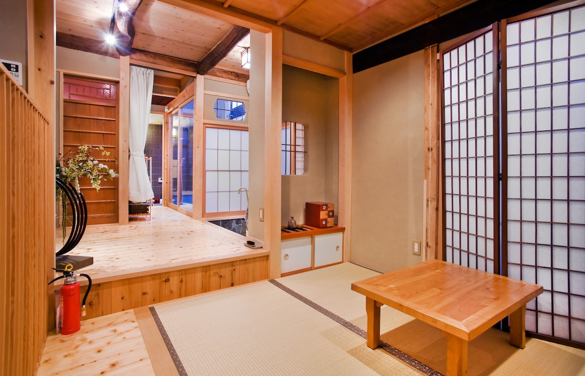 A traditional Japanese interior showcases wooden beams and tatami flooring. A low wooden table sits centered in the room, with sliding shoji doors in the background, allowing natural light to filter through. The space offers a cozy and calming atmosphere, enhanced by natural materials.