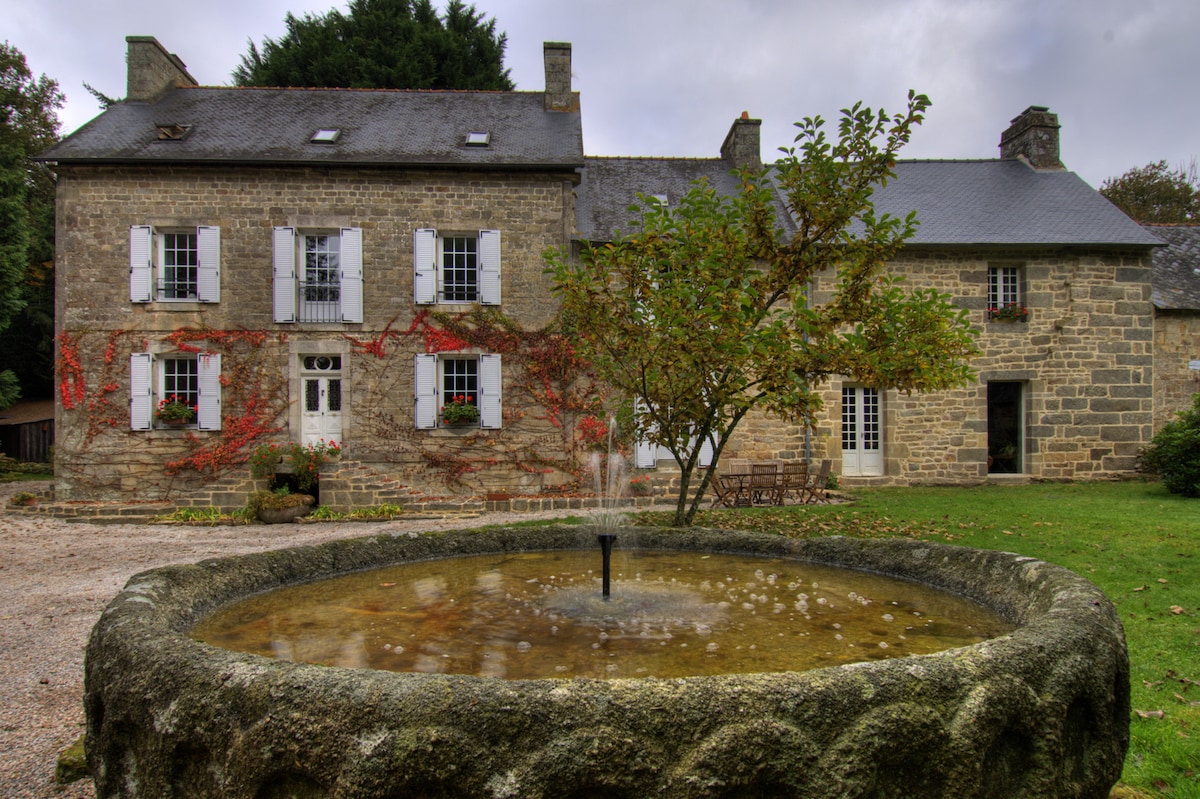 The historic manor features a stone façade, complemented by white shutters and climbing vines. A rustic stone fountain is located in the foreground, surrounded by a landscaped area. The house has large windows, allowing natural light to illuminate the inviting exterior.