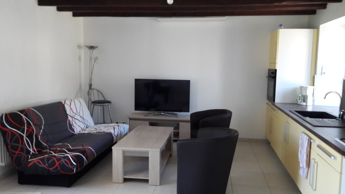 The living area features a comfortable seating arrangement with a patterned sofa and two black armchairs. A wooden coffee table sits in front of the sofa, while a smart TV is positioned against the wall. Bright cabinets in the kitchen area enhance the space.