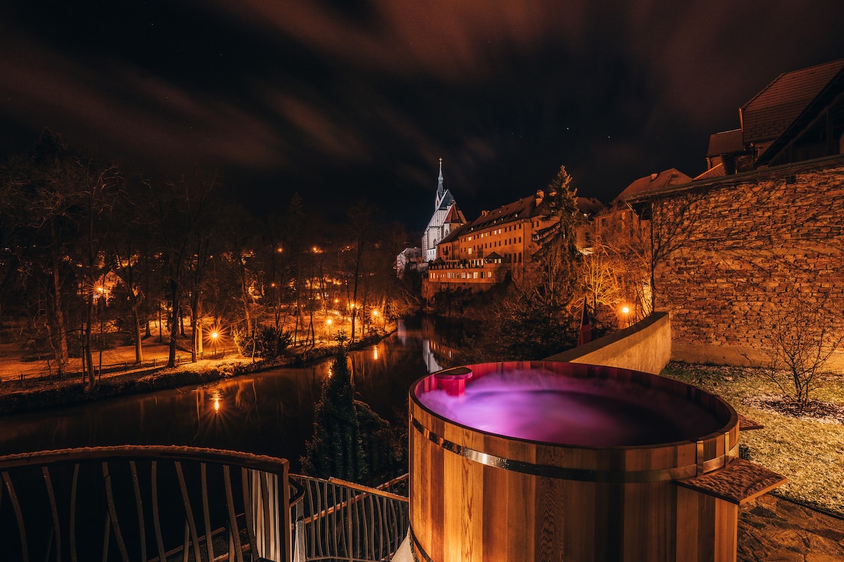 An outdoor jacuzzi is positioned on a terrace, surrounded by wooden decking. Soft purple lighting reflects off the water, creating a serene atmosphere. In the background, illuminated buildings and trees line the riverbank, enhancing the tranquil view under a night sky.