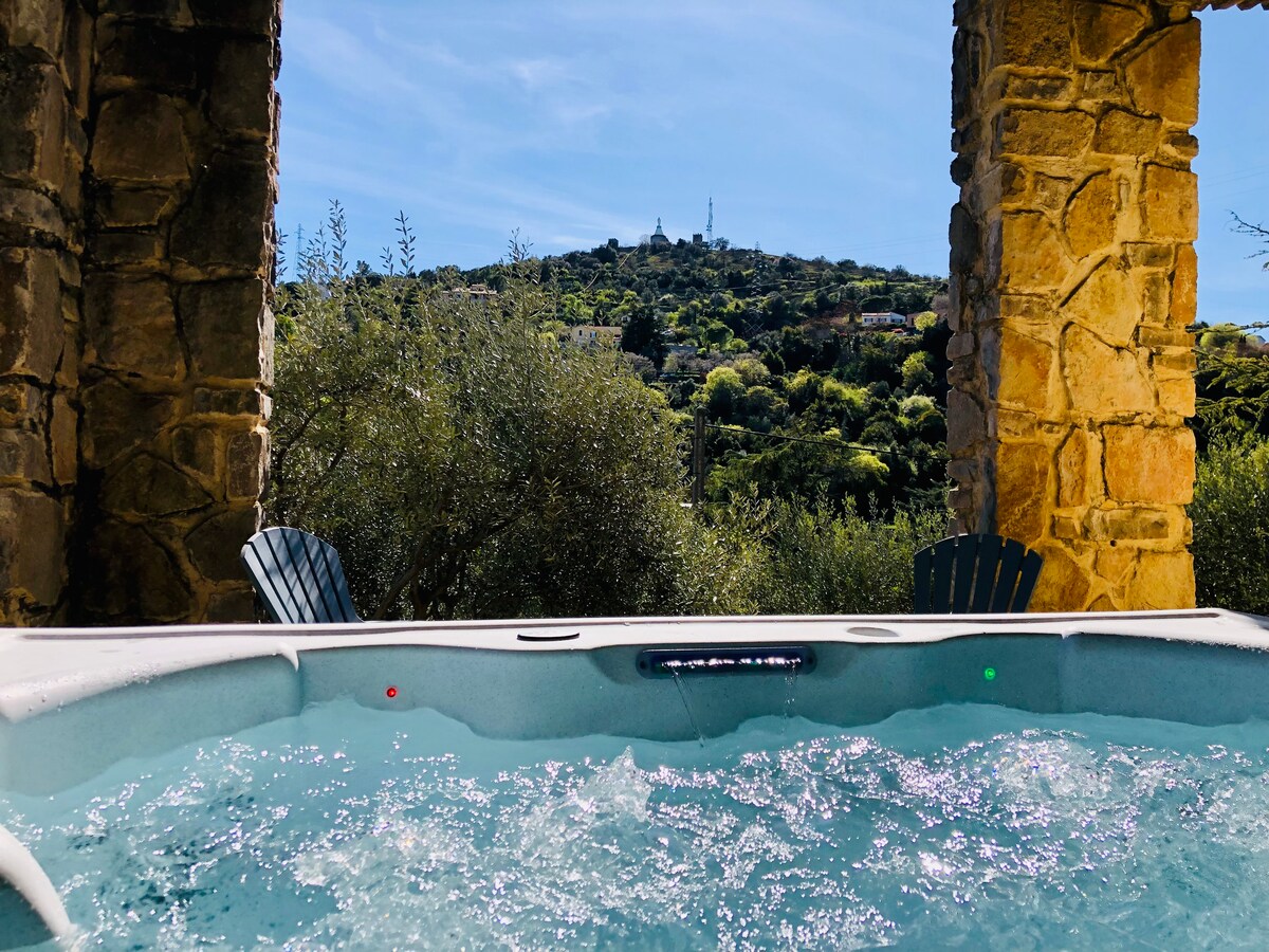 A jacuzzi is positioned under stone archways, overlooking a hillside landscape. Olive trees surround the area, and the sky is bright with a few clouds. Two wooden adirondack chairs are visible nearby, inviting relaxation amid the scenic backdrop.