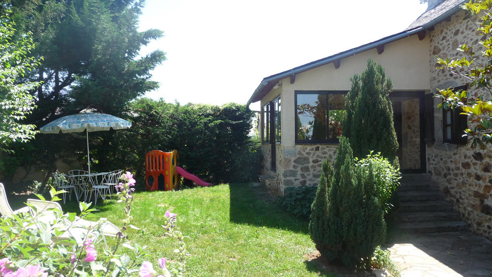 A charming stone exterior of the house is visible, surrounded by a green lawn. A playset with a slide stands beside colorful patio furniture under a shaded umbrella. Lush bushes and blooming flowers enhance the outdoor space, providing a welcoming atmosphere.