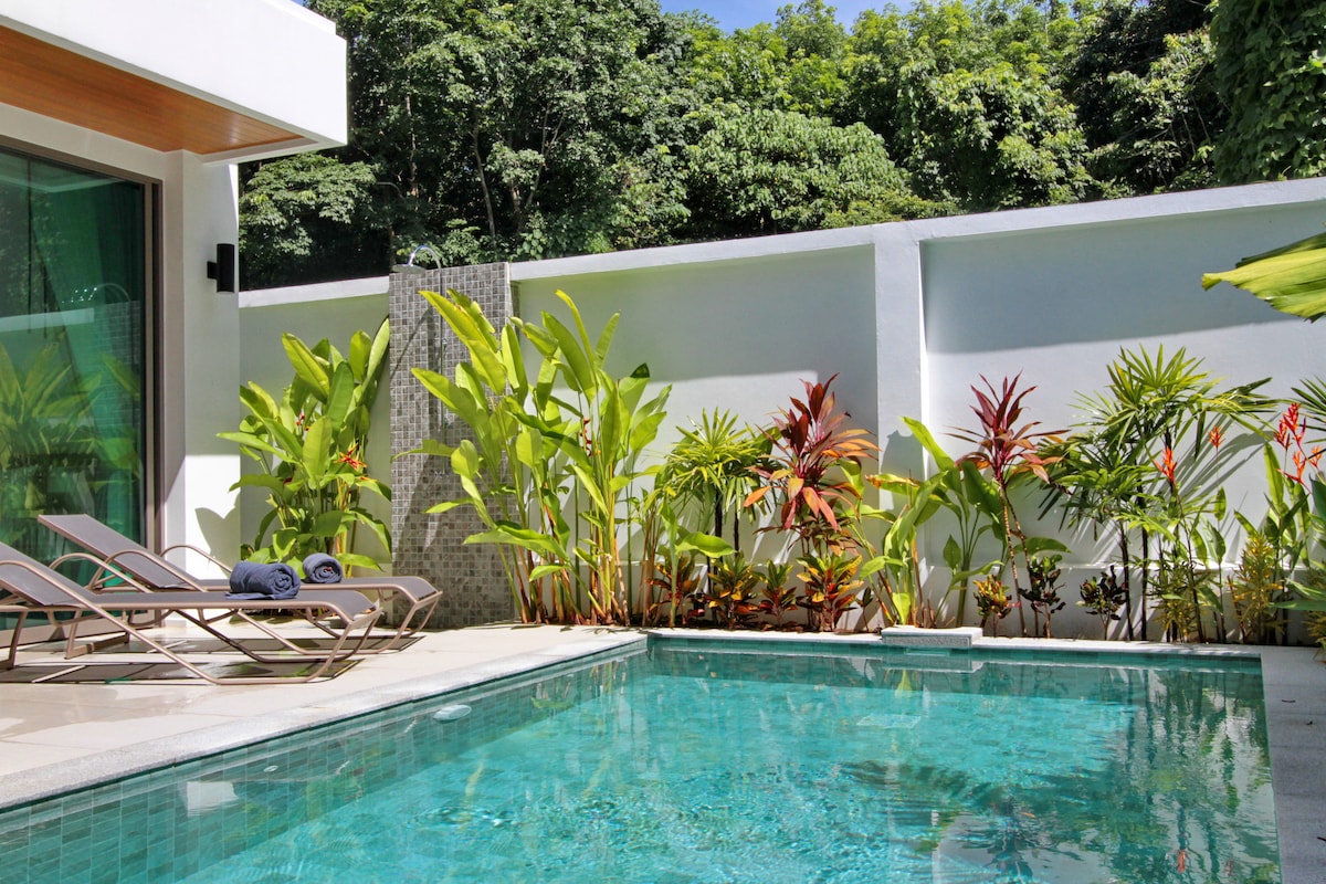 A private pool area is surrounded by lush tropical greenery, featuring various palm and flowering plants. Comfortable lounge chairs are positioned beside the crystal-clear water, creating a serene outdoor setting. Natural light highlights the clean lines of the modern architecture and the inviting pool.