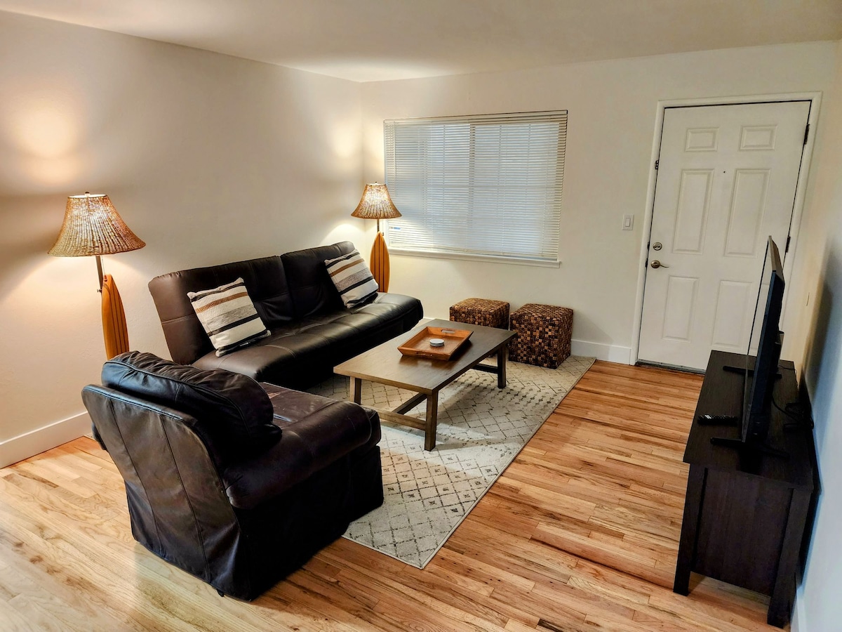 A living area features a dark leather sofa accompanied by two decorative lamps. A coffee table sits centrally, positioned over an area rug. Additional seating includes textured ottomans, while a flat-screen television is mounted on the wall. Natural light filters through a window beside the entrance door.
