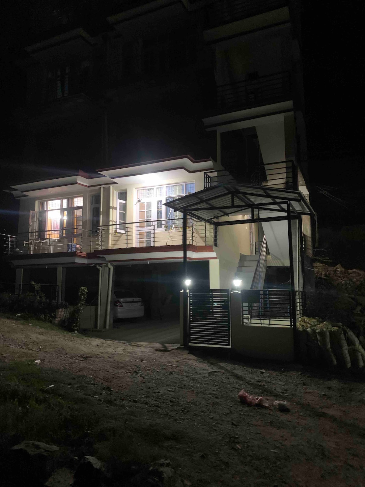 The exterior of the property is shown at night, featuring bright lights illuminating the entrance. A covered area is visible, providing shelter above the steps. A private parking space is included, with a vehicle parked nearby. The overall architecture is modern and welcoming.