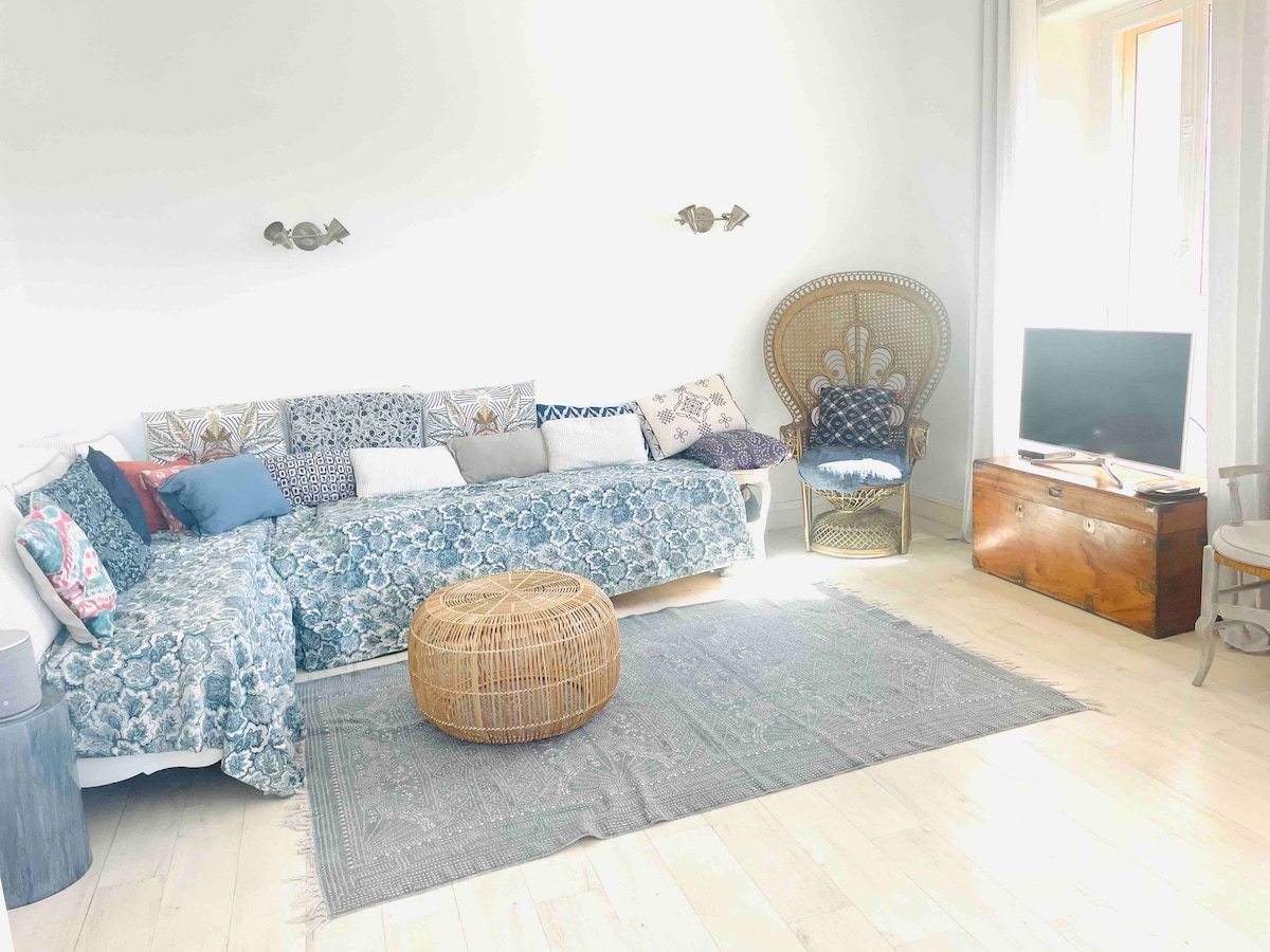 A spacious living area features a light-colored floor and soft natural light from large windows. A patterned sofa is complemented by decorative pillows and a round woven ottoman. A wooden TV stand holds a flat-screen television, while a rattan chair provides additional seating.