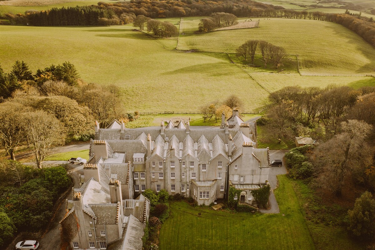 An aerial view reveals the sprawling Dunskey Castle surrounded by lush, rolling hills. The landscape showcases a patchwork of greenery, with trees dotting the property and paths leading away into the distance, creating a serene and inviting scene.