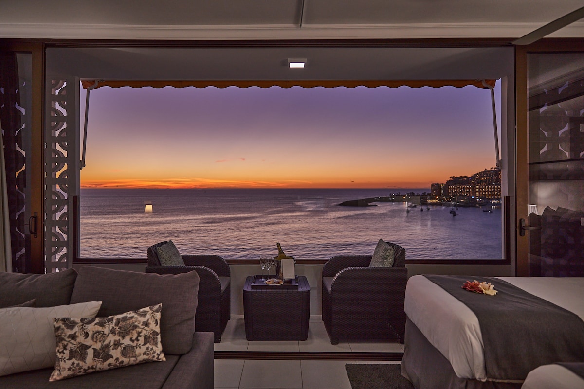 A panoramic view of the sunset over the sea is framed by large windows. The tranquil scene is complemented by a cozy seating area with dark wicker furniture and soft cushions. A bottle of champagne and a plate of fruit are elegantly arranged on a low table.