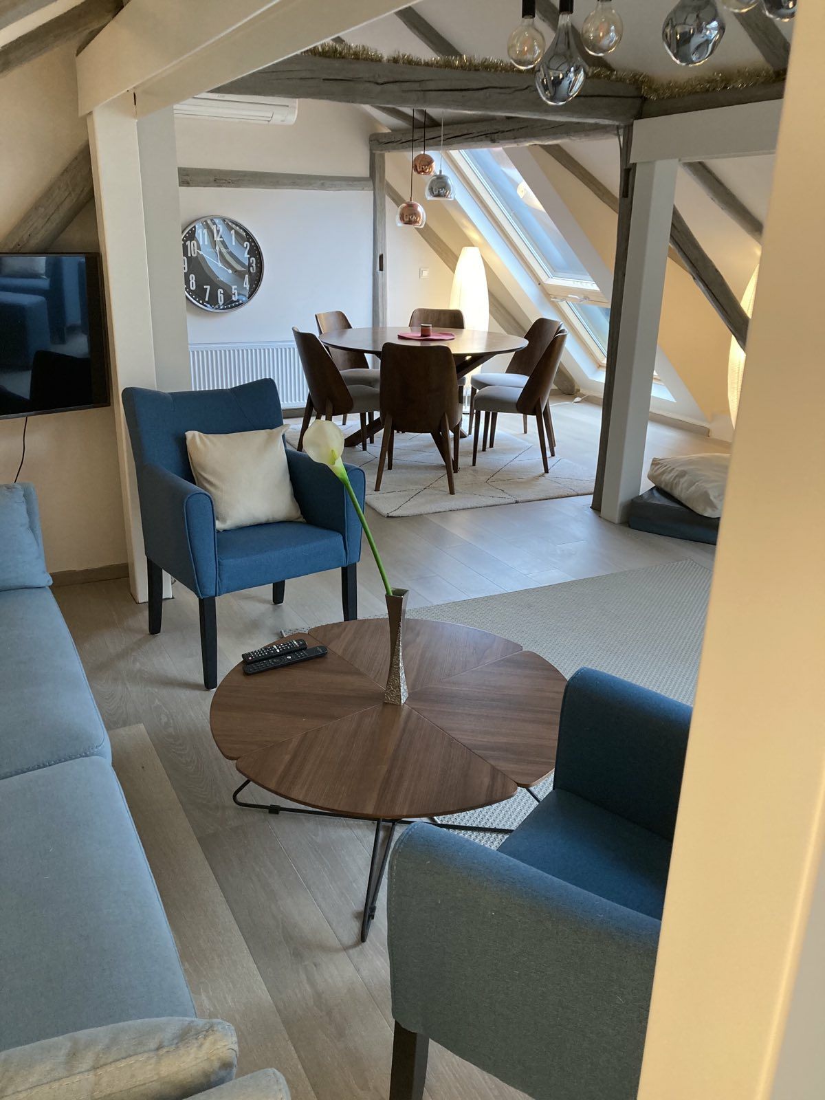 A spacious living area features two light blue armchairs facing a round wooden coffee table with a single flower vase. A sleek television is mounted on the wall, and the dining area is visible in the background, furnished with a dark wooden table and chairs. Skylight windows contribute to a bright atmosphere.