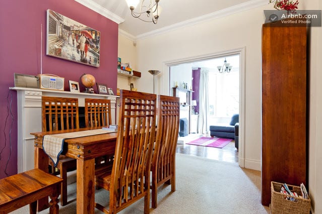 A dining area features a wooden table surrounded by multiple chairs, set against a vibrant purple wall. A small side table holds items and a framed artwork is displayed above the mantel. Soft light fills the space from an adjacent living area, enhancing the open feel.