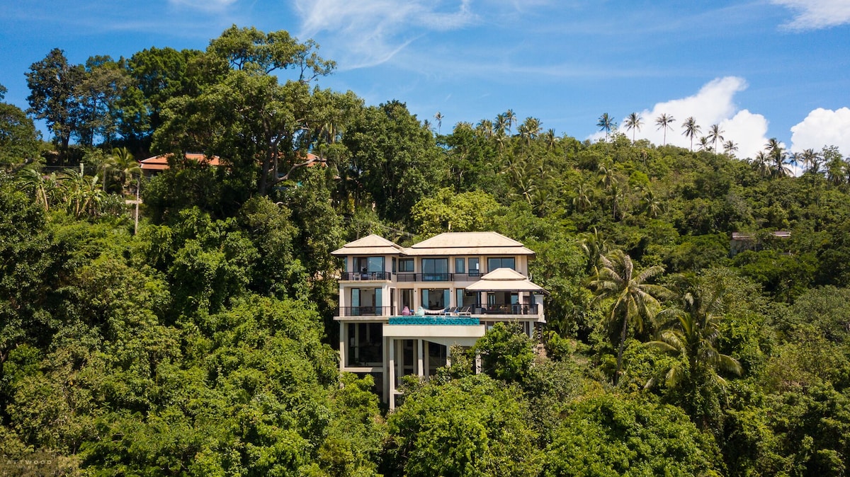 The villa is situated on a hillside, surrounded by lush greenery and palm trees. Its three-story structure features a rooftop terrace and a pool area that overlooks the tropical landscape, providing a sense of privacy and tranquility amidst the natural surroundings.