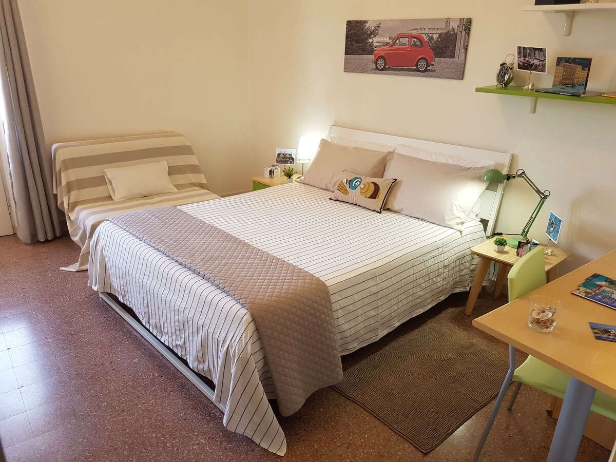 A spacious bedroom features a large double bed with a striped coverlet and a plush decorative pillow. A desk with a lamp and a small seating area are included. Natural light enters through a window, illuminating the modern furnishings and warm tones in the room.