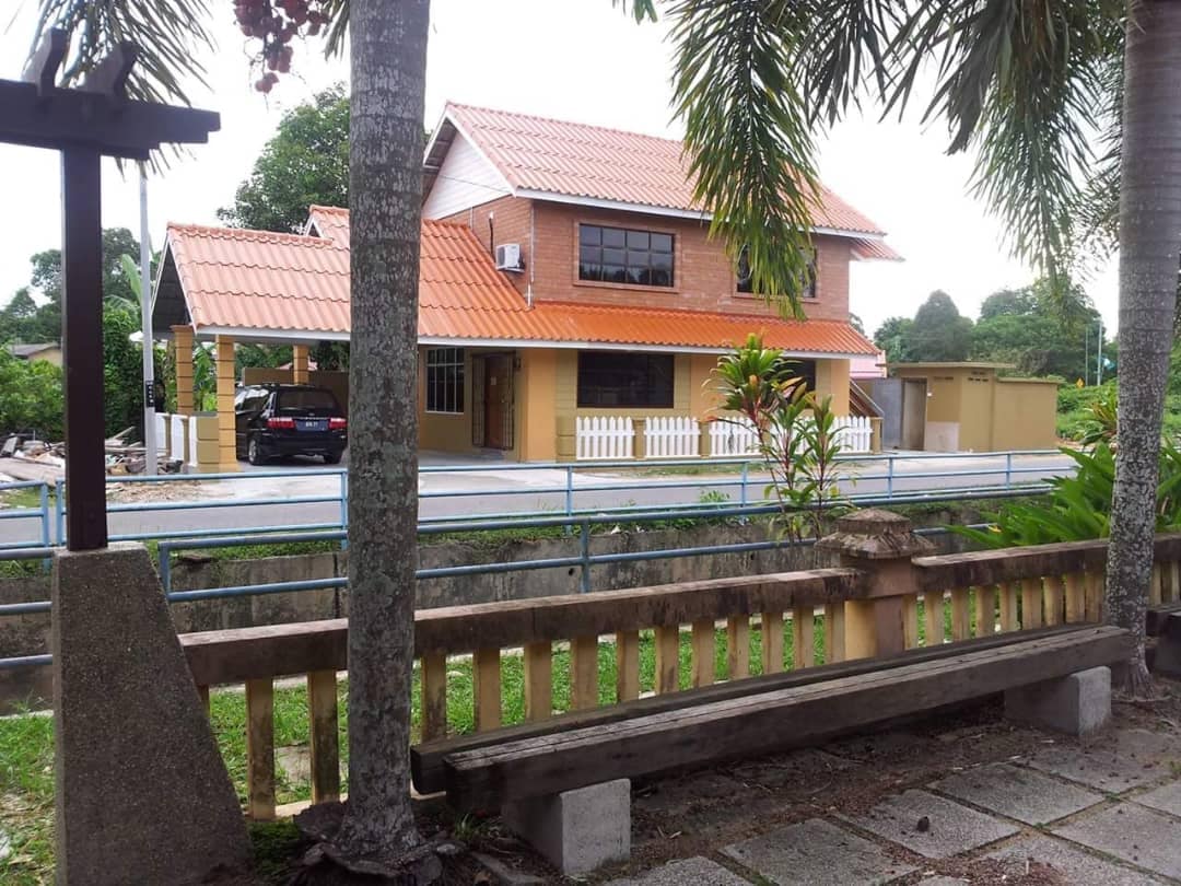 A two-story house is framed by palm trees and a wooden bench in the foreground. The building features a red tile roof, large windows, and a driveway with a parked car. A white fence borders the property, enhancing the welcoming outdoor space.