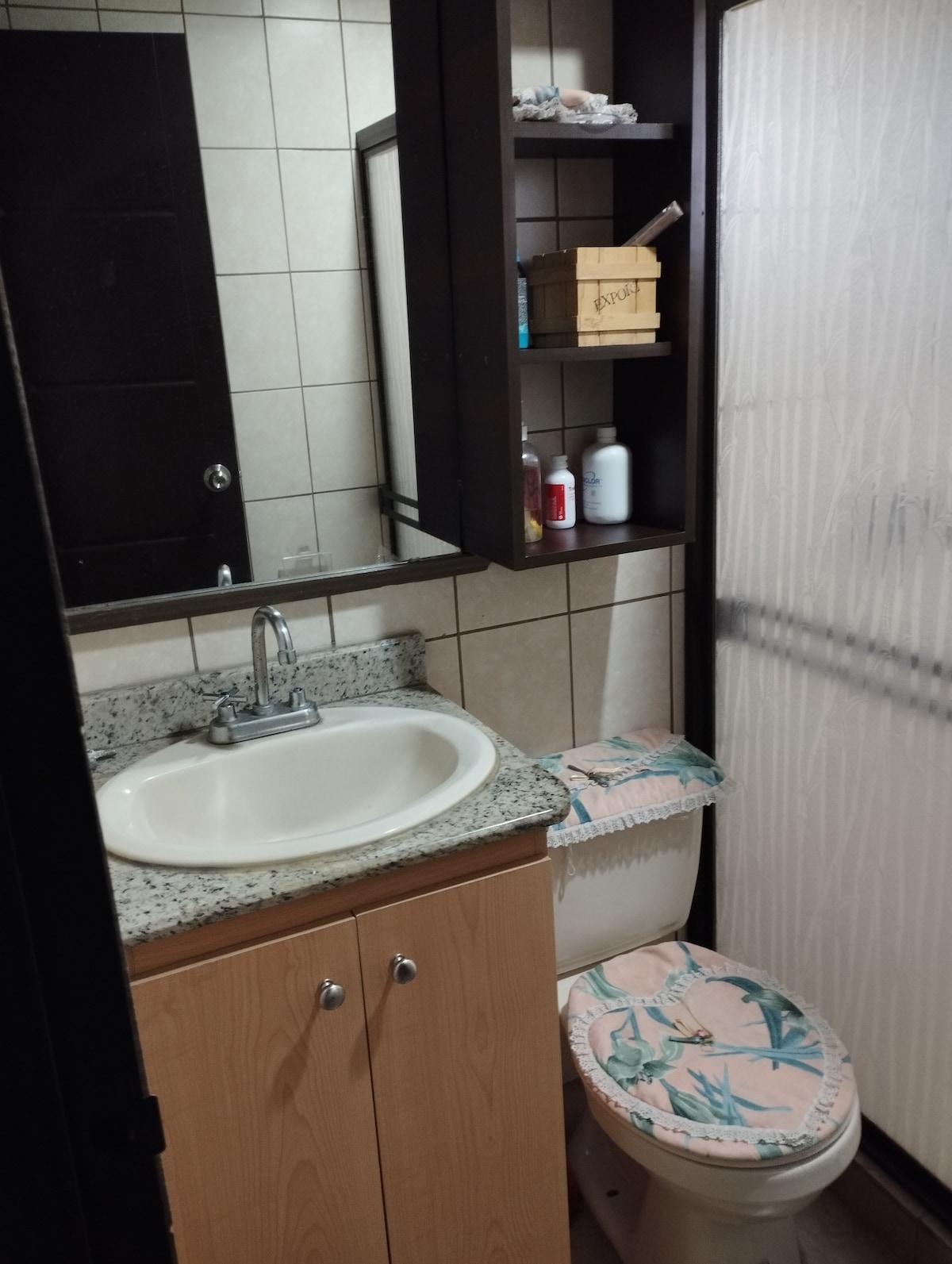 The bathroom features a granite countertop with a sink and storage cabinet below. A mirror reflects the space while toiletries are stored in a wooden box on a shelf. A shower is enclosed with frosted glass, and a patterned cover adorns the toilet.